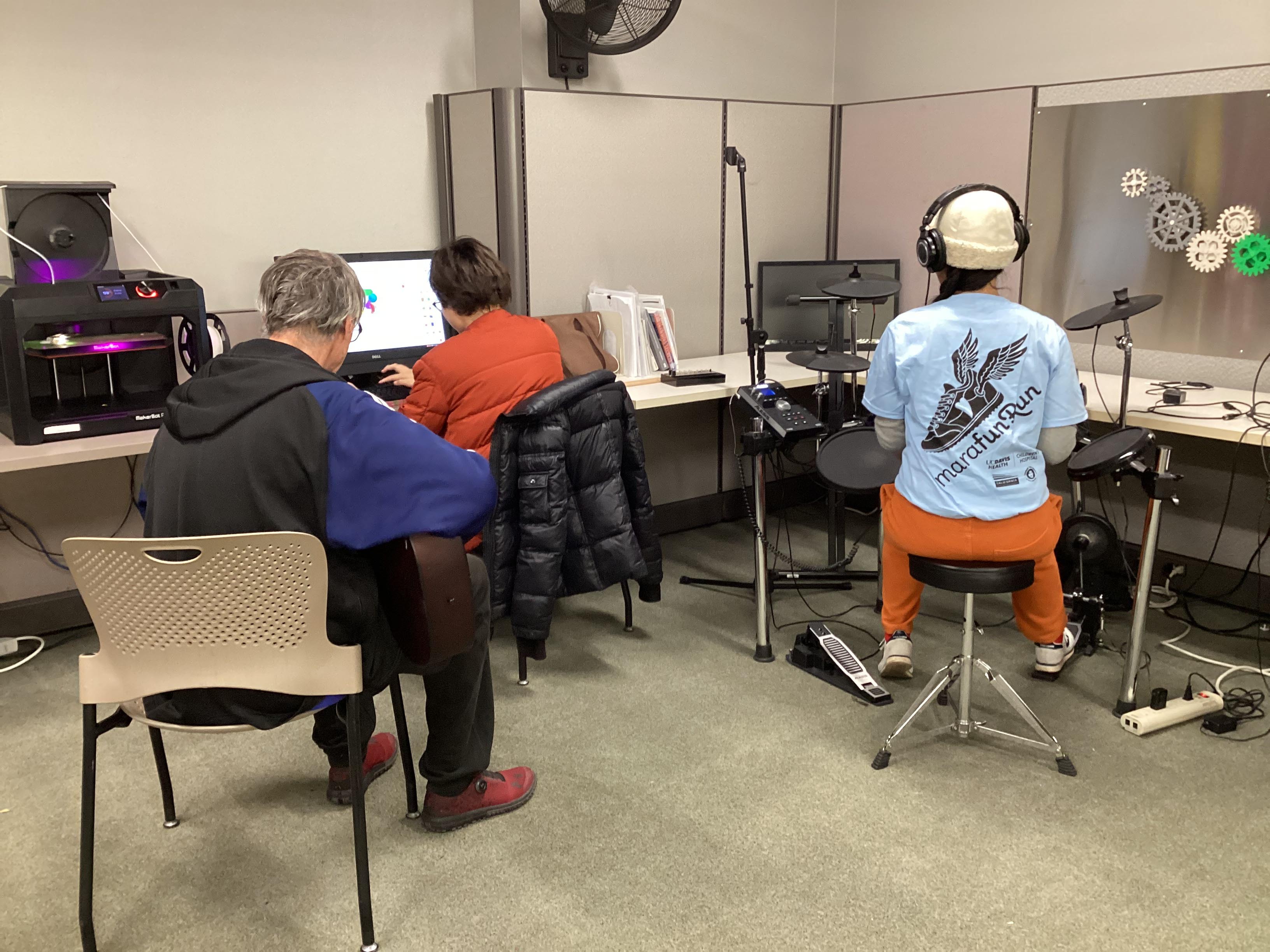 Musicians play the guitar and drums inside the Makerspace at the Central Library. Another person sits at the desk with a computer and mixer to record the track. There is a 3D printer on the desk nearby.