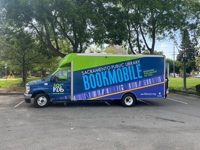 The Library's new, all-electric vehicle is parked in front of a city park. The vehicle's graphics are bright green, dark blue and purple, and with words Sacramento Public Library Bookmobile, bringing the library to you."