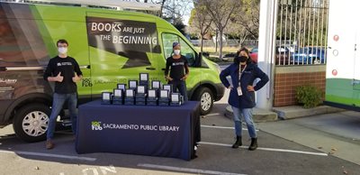 Three library staff members stand in front of a green and black library van with a table full of Wi-Fi hotspots.