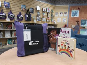 On a table inside of a library sits a black and purple tote bag with a white tag that says “Patrones.” Next to the bag are two books: “de Colores” and “Amor de pelo.”