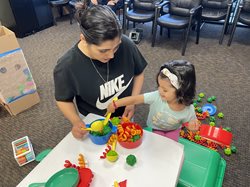 A child with short dark hair and a white headband plays with colorful toys on a white table. The child’s caregiver, in a black and yellow shirt, their dark hair up, kneels down to play with the child.