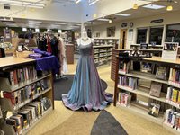 Formal attire, including a long purple and blue dress on a mannequin, arranged between library book shelves.