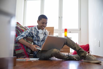 A smiling teen wearing a blue plaid flannel shirt, jeans and sneakers and white headphones sits on the floor by their bed with a silver laptop on their lap.