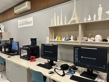 A 3D printer and several computers sit side-by-side on a long desk. Shelves above display items like the Eiffel Tower, a skull and figures created on a 3D printer at the Design Spot in the Arcade Library.