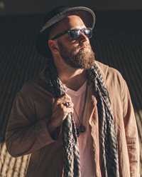Sacramento Poet Laureate Andru Defeye wears a black brimmed hat and dark sunglasses while looking up and away from the camera. He is wearing a light colored V neck shirt, a tan jacket, 2 necklaces, and an untied scarf around his neck. One hand is grasping the scarf and has a purple ring on it.