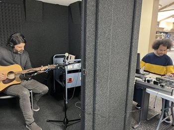 A musician plays guitar inside of a sound booth at the Martin Luther King, Jr. Library. Another person sits at the computer and sound mixer outside to record the track.